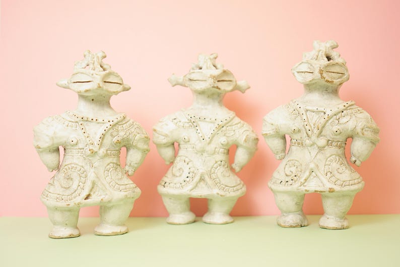 May include: Three off-white ceramic figurines with detailed facial features and ornate clothing. The figures stand with their arms at their sides, and have a unique, stylized appearance. The background is a soft pink and green.