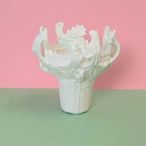 May include: A white, decorative vase with an intricate, textured design. The vase features a flared top with sculpted elements and a cylindrical base. The background is split between pink and green.