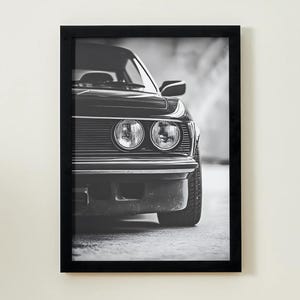 Könnte beinhalten: Schwarz-Weiß-Druck in einem Rahmen, der ein klassisches Auto zeigt. Das Bild konzentriert sich auf die Front des Autos, mit Scheinwerfern, Kühlergrill und einem Teil des Reifens. Der verschwommene Hintergrund erzeugt Tiefe.