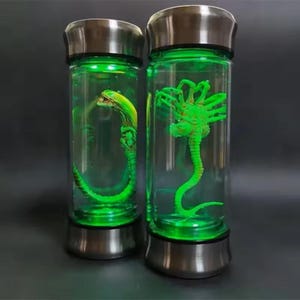 May include: Two clear glass tumblers with stainless steel lids and bases, each containing a green alien figure. The tumblers are illuminated with green LED lights, creating a sci-fi or horror theme.