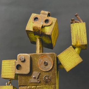 May include: A handcrafted, wooden robot sculpture with a vintage aesthetic. The robot features a box-shaped head and body, with articulated arms. The color palette includes shades of yellow, brown, and metallic accents, giving it a steampunk vibe. The robot's arm is raised.