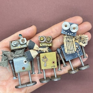 May include: Three small wooden robots with different colored bodies and faces. The robots have metal legs and arms, and some have additional details like keys and fabric.
