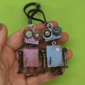 May include: Two handmade robot pendants. One is lavender and the other is light blue. Both have metal bolt arms and legs, and metal screw eyes. Each robot has a small heart painted on the front. They hang from a black cord.