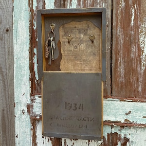 Può includere: Un portachiavi da parete rettangolare in legno con cornice grigio scuro. La sezione superiore presenta uno sfondo con un disegno architettonico vintage e tre ganci in metallo, uno dei quali sostiene un mazzo di chiavi. Il pannello inferiore riporta 1934.