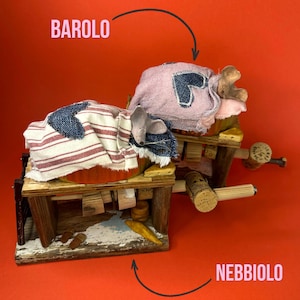 May include: A whimsical diorama featuring two miniature mice resting on wooden structures. One mouse is under a striped blanket, the other under a pink cloth with a heart. The word "BAROLO" and "NEBBIOLO" are written above and below the mice.