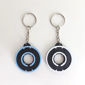 May include: Two keychains with circular black and blue or white designs. Each keychain has a silver chain and ring. The circular design has a black center with a light blue or white outer ring and inner circle.