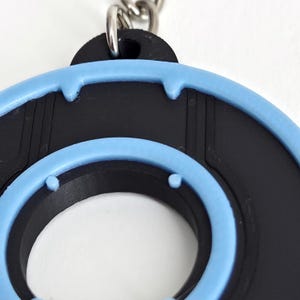 May include: Close-up of a black and light blue keychain. The keychain has a circular design with a light blue border and a black interior. The keychain is attached to a silver chain.