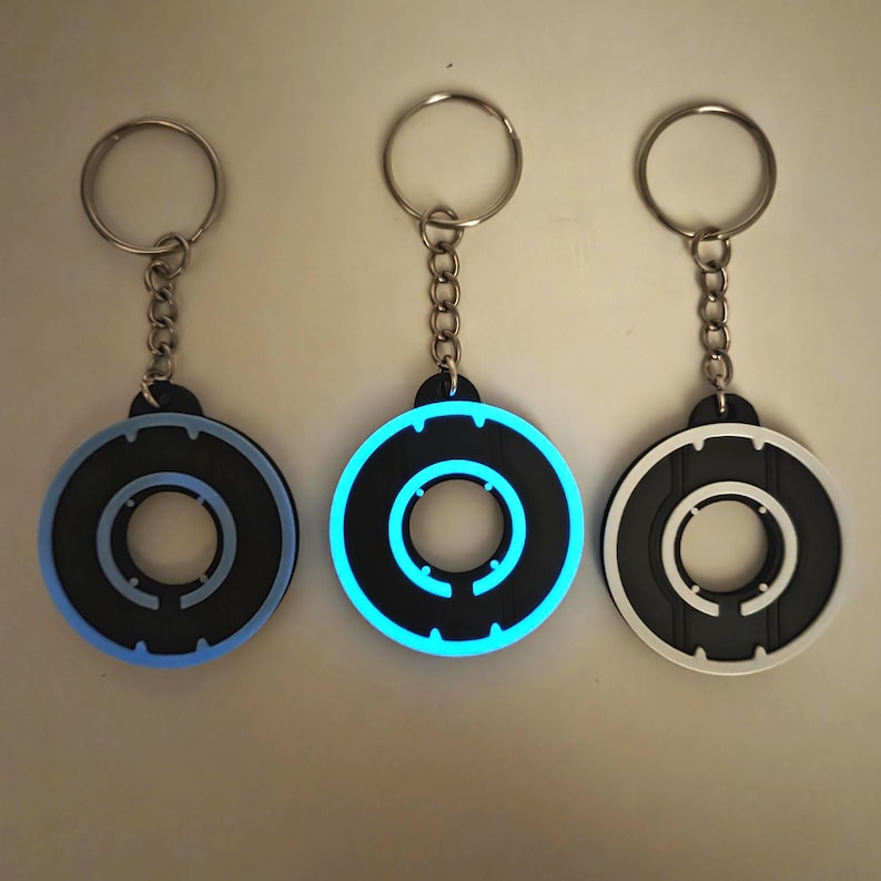 May include: Three keychains with circular, black and blue designs. The keychains have a silver chain and ring. One keychain glows blue, another has a blue outline, and the third has a white outline. The keychains are likely made of rubber or plastic.