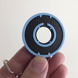 May include: A black and light blue circular keychain with a key ring. The keychain has a light blue outer ring and a black inner circle with a white center. The design features vertical lines and small circles.