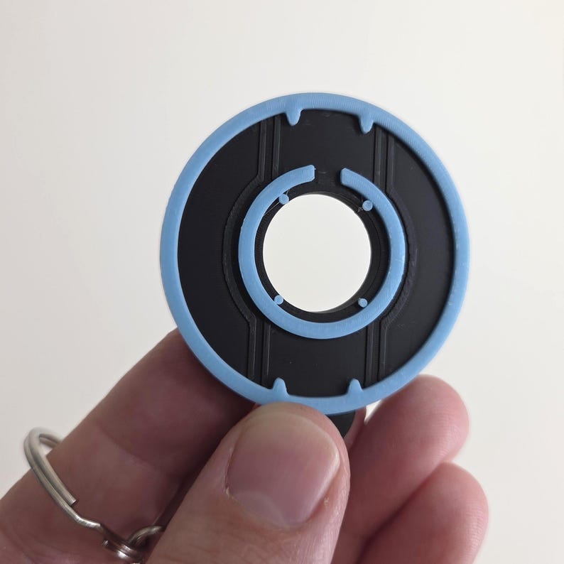 May include: A black and light blue circular keychain with a key ring. The keychain has a light blue outer ring and a black inner circle with a white center. The design features vertical lines and small circles.