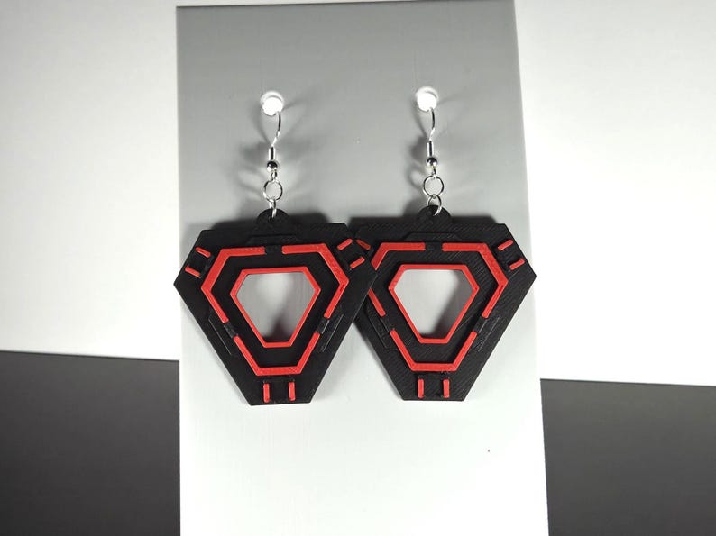 May include: A pair of black and red geometric earrings. Each earring features a black, diamond-shaped frame with a red, hexagonal design in the center. The earrings hang from silver-colored hooks.
