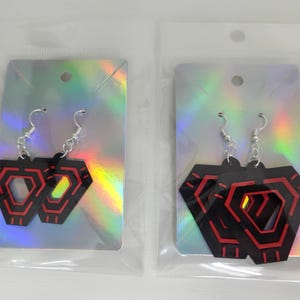 May include: Two pairs of black and red geometric earrings with silver hooks. The earrings are in clear plastic packaging, displayed on iridescent silver cards. The earrings have a layered design with red accents.