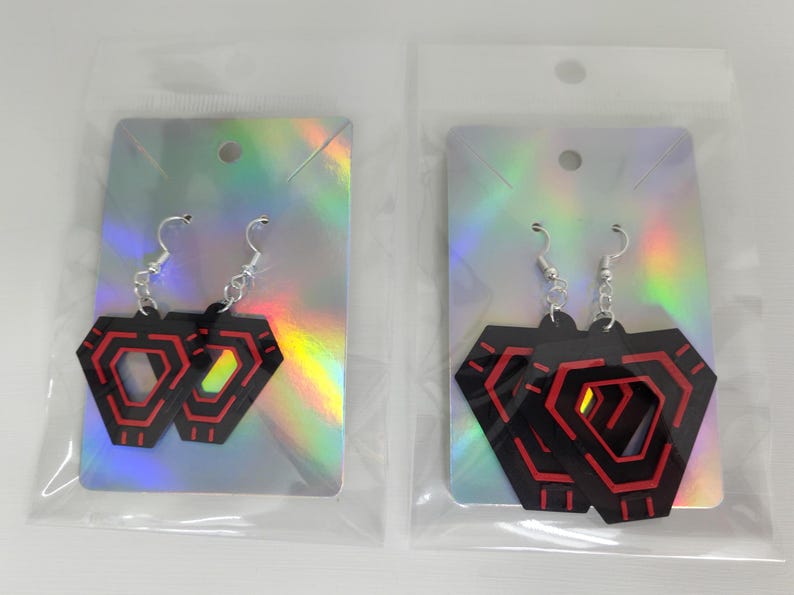 May include: Two pairs of black and red geometric earrings with silver hooks. The earrings are in clear plastic packaging, displayed on iridescent silver cards. The earrings have a layered design with red accents.