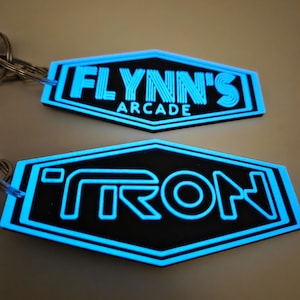 May include: Two black and blue keyrings. The top keyring reads "FLYNN'S ARCADE" in a glowing blue font. The bottom keyring reads "TRON" in a similar glowing blue font. Both keyrings have a hexagonal shape.