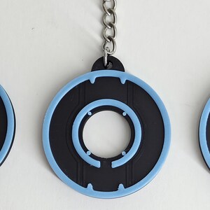 May include: Black and light blue circular keychains with a silver chain. The keychains have a black center with a light blue outline and details. The design features a circular shape with a central hole and a series of smaller circles.