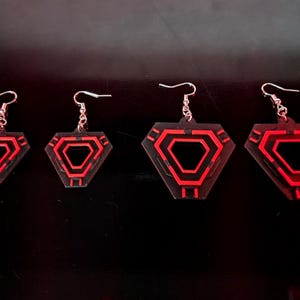 May include: Four pairs of geometric earrings with a red and black design. The earrings are diamond-shaped with a red outline and a red interior design. The earrings are suspended from silver-colored hooks.