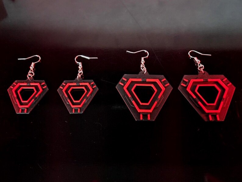 May include: Four pairs of geometric earrings with a red and black design. The earrings are diamond-shaped with a red outline and a red interior design. The earrings are suspended from silver-colored hooks.