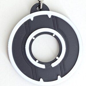 May include: A black and white circular keychain with a unique design. The keychain features a dark gray center with a white outer ring and inner circle. The design includes small white dots and lines, and a silver metal ring for attachment.