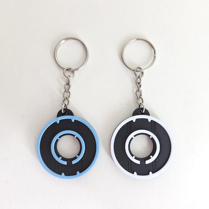 May include: Two keychains with circular black and blue or white designs. Each keychain has a silver chain and ring. The circular design has a black center with a light blue or white outer ring and inner circle.