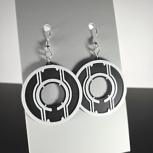 May include: A pair of black and white circular earrings with a geometric design. The earrings are made of a black material with white accents and are suspended from silver-colored hooks. The earrings are displayed on a gray card.
