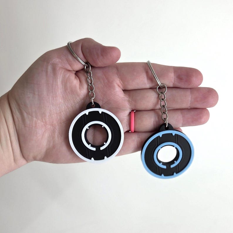 May include: Two circular keychains with a black and white design and a black and light blue design. Each keychain has a silver chain and ring. The keychains are held in a hand.