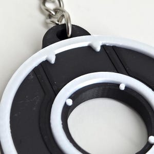 May include: A black and white circular keychain with a silver chain. The keychain has a black center with a white outer ring and accents. The design is reminiscent of a retro video game aesthetic.
