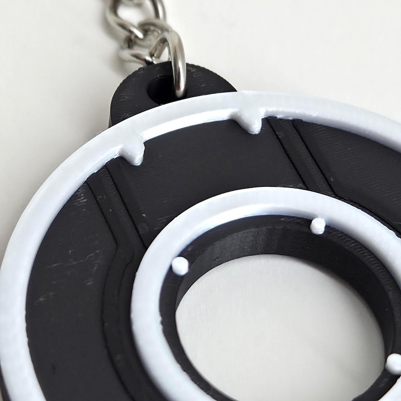 May include: A black and white circular keychain with a silver chain. The keychain has a black center with a white outer ring and accents. The design is reminiscent of a retro video game aesthetic.