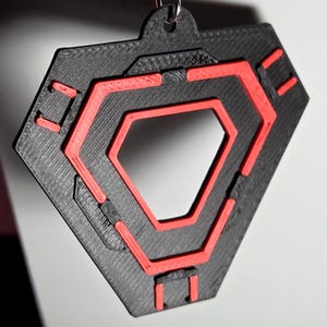 May include: A black and red geometric pendant with a hexagonal design. The pendant has a textured surface and a small loop at the top for hanging. The red accents create a striking contrast against the black.