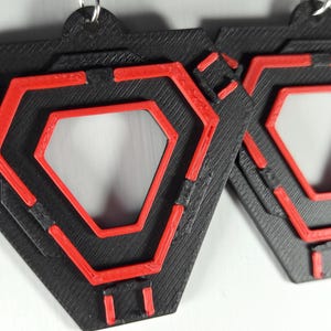 May include: Pair of geometric earrings. Each earring is a black, layered, hexagonal shape with a red, open-center hexagon design. The earrings have a textured appearance and are suspended from silver-colored hooks.