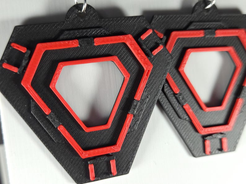 May include: Pair of geometric earrings. Each earring is a black, layered, hexagonal shape with a red, open-center hexagon design. The earrings have a textured appearance and are suspended from silver-colored hooks.
