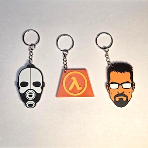 May include: Three keyrings with silver rings and chains. One features a black and white gas mask design. Another has an orange and yellow triangle with a lambda symbol. The third keyring has an orange and black face.