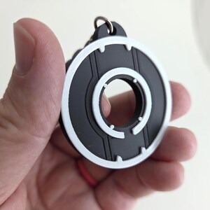 May include: A black and white circular keychain with a silver key ring. The design features a series of concentric circles with white accents. The keychain is held in a hand.