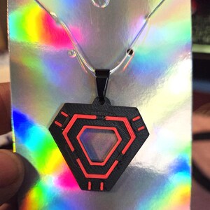 May include: A black pendant necklace with a geometric design featuring red accents. The pendant is shaped like a hexagon with a central opening and hangs from a silver chain. The background is a holographic card.