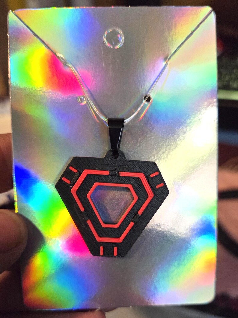 May include: A black pendant necklace with a geometric design featuring red accents. The pendant is shaped like a hexagon with a central opening and hangs from a silver chain. The background is a holographic card.