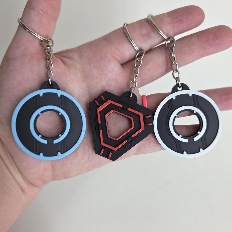 May include: Three keychains with silver rings and chains. The keychains have black and colored designs. One is a black circle with a light blue outline, another is a black and red geometric shape, and the last is a black circle with a white outline.
