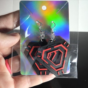 May include: Black and red geometric earrings with silver-colored hooks. The earrings are in a clear plastic package on a holographic display card. The design features a series of red lines on a black background.