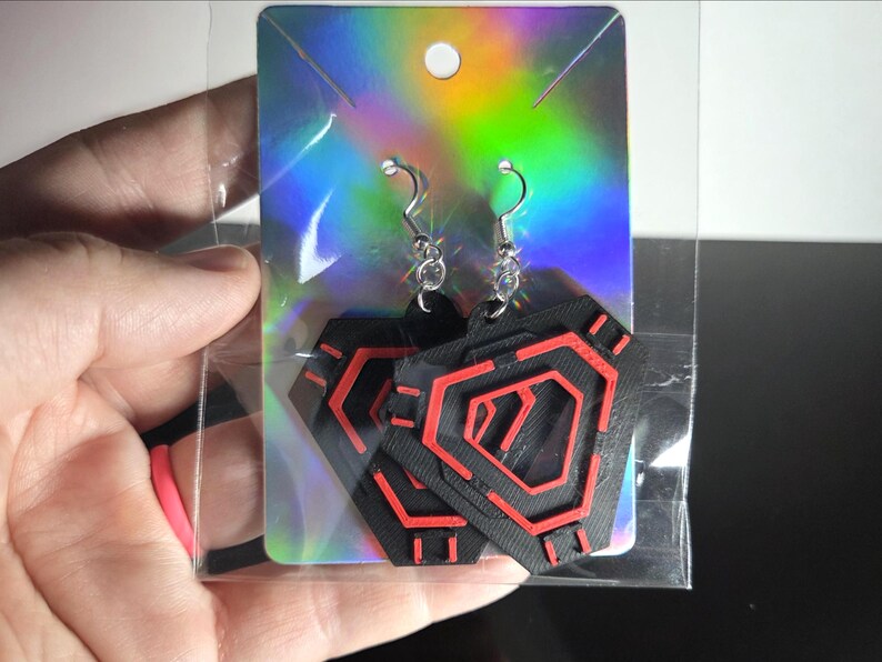 May include: Black and red geometric earrings with silver-colored hooks. The earrings are in a clear plastic package on a holographic display card. The design features a series of red lines on a black background.