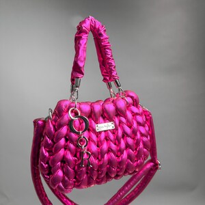 May include: A vibrant fuchsia handbag with a braided design. The bag features a top handle and a detachable shoulder strap. A silver charm with the letter "R" hangs from the handle. A small silver tag is attached to the bag.