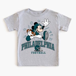 A gray t-shirt with a graphic design featuring a cartoon character in a football uniform, along with various football-related icons and text.