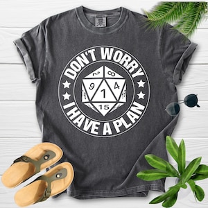 May include: Dark gray t-shirt with the text "DON'T WORRY I HAVE A PLAN" in a white circular design. The design includes a 20-sided die. The shirt is paired with sandals and sunglasses.