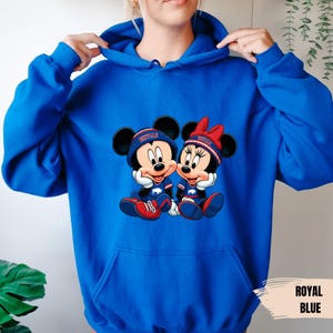 A person wearing a blue hoodie with a graphic of two cartoon mice, one wearing a baseball cap and the other a bow, sitting together.