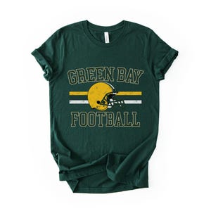 May include: Dark green t-shirt with the words "GREEN BAY FOOTBALL" in a vintage font. A yellow football helmet is centered between the words, with a yellow and white stripe design. The shirt is made of a soft material.