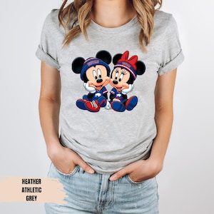 A woman wearing a grey t-shirt with a graphic of two cartoon mice, one wearing a blue hat and the other a red hat, sitting together.