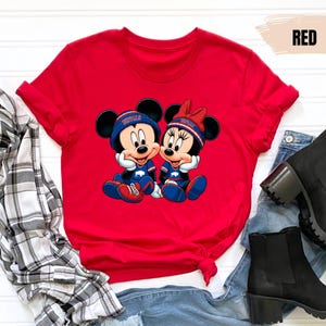 A red t-shirt with a cartoon image of Minnie Mouse and Mickey Mouse, along with a plaid shirt, jeans, and black boots, all laid out on a white background.
