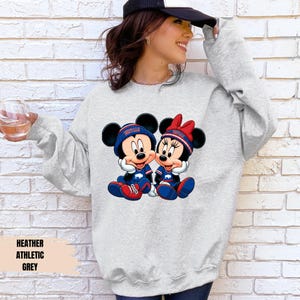 A woman wearing a grey sweatshirt with a graphic of Minnie Mouse and Mickey Mouse, holding a drink and smiling.