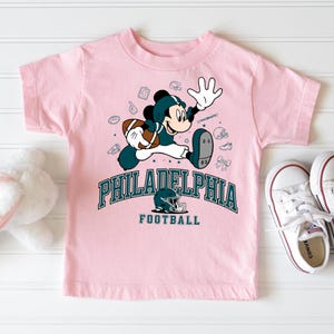 A pink t-shirt with a cartoon character and text related to Philadelphia football, along with a pair of white sneakers and a stuffed animal.