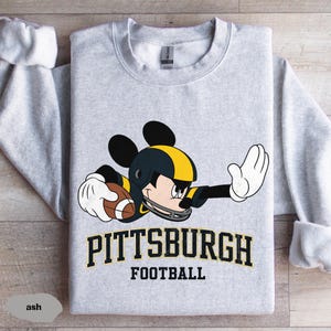 May include: Ash-colored sweatshirt featuring a cartoon Mickey Mouse in a football uniform. The design includes a black and yellow helmet, holding a football, with the words "Pittsburgh Football" in black and gold.