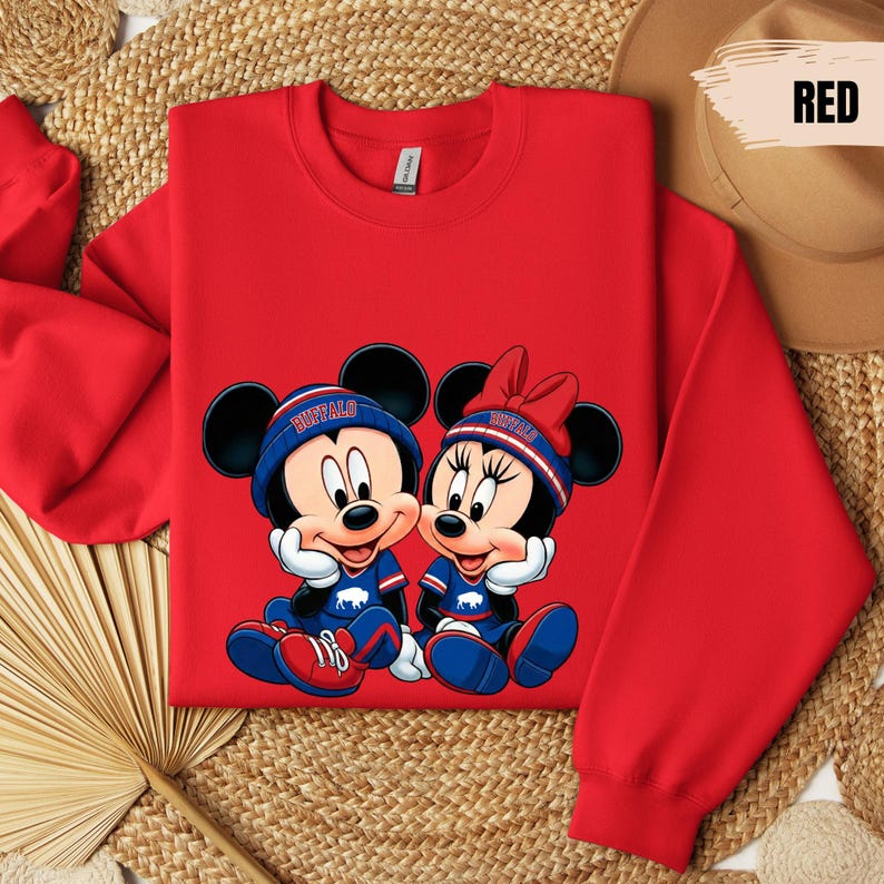 A red sweatshirt with a cartoon image of Minnie Mouse and Mickey Mouse, along with a red hat, is displayed on a woven mat.