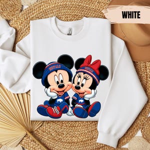 A white long-sleeved shirt with a graphic of two cartoon mice, Minnie Mouse and Mickey Mouse, sitting together. The mice are wearing matching blue and red outfits. The shirt is laid out on a woven mat, and there is a hat visible in the background.