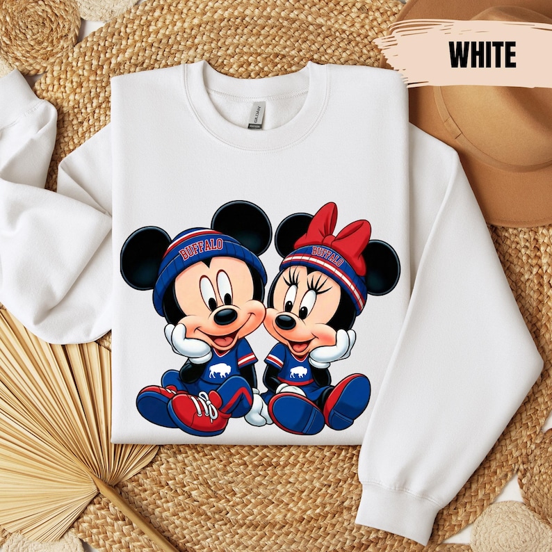 A white long-sleeved shirt with a graphic of two cartoon mice, Minnie Mouse and Mickey Mouse, sitting together. The mice are wearing matching blue and red outfits. The shirt is laid out on a woven mat, and there is a hat visible in the background.
